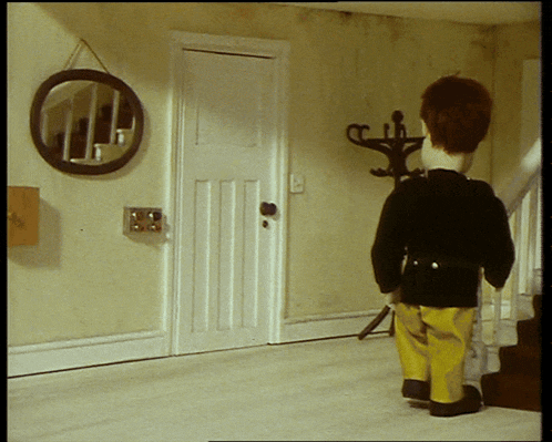 a doll in a black jacket and yellow pants stands in a hallway