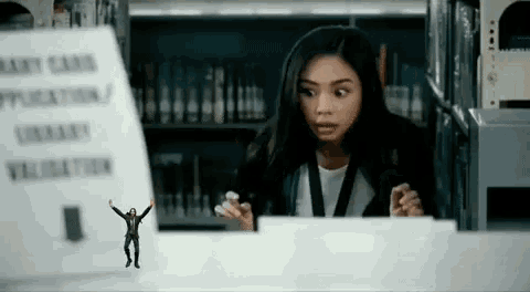a woman is sitting at a desk in a library with a miniature man standing next to her .