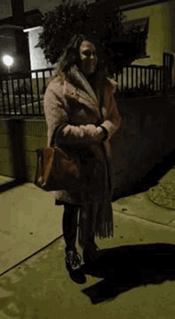 a woman is standing on a sidewalk at night holding a brown purse .