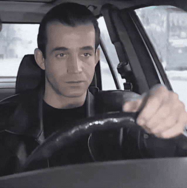 a man in a black jacket is driving a car and looking at the camera