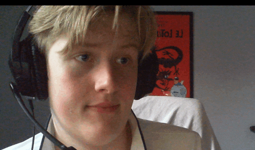 a young man wearing headphones and a microphone looks at the camera in front of a red poster that says totoro