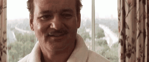 a man with a mustache is smiling in front of a window while wearing a white robe .