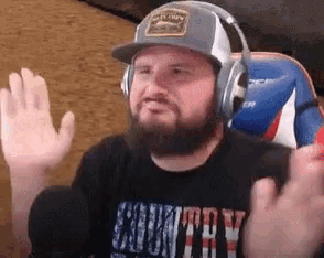 a man with a beard wearing headphones and a hat is waving his hands in front of a microphone .