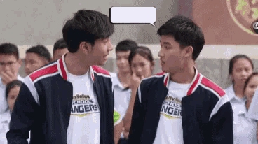 two young men are standing next to each other in front of a crowd of people and talking to each other .