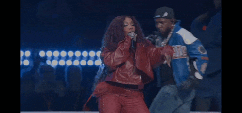 a woman in a red jacket singing into a microphone while a man in a blue jacket stands behind her