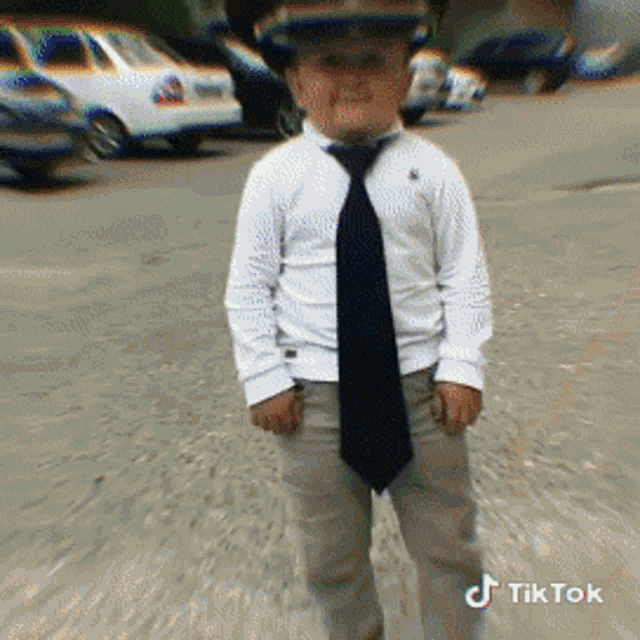 a young boy wearing a hat and tie is standing in a parking lot with cars in the background