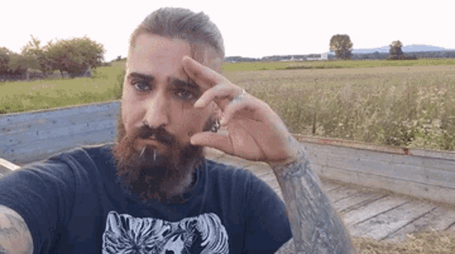 a man with a beard and a tattoo on his arm is taking a selfie in front of a field .