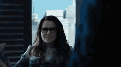 a woman wearing glasses is standing in front of a window in a dark room .