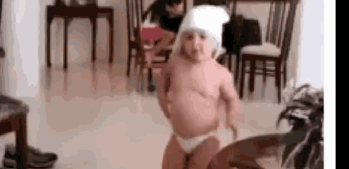 a baby is dancing in a living room while wearing a diaper and a towel on his head .