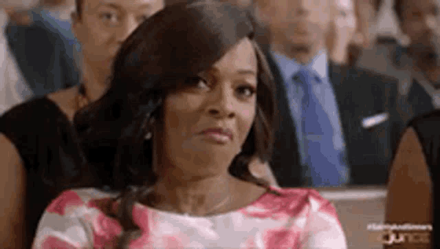 a woman in a pink and white dress is sitting in a crowd of people making a funny face .