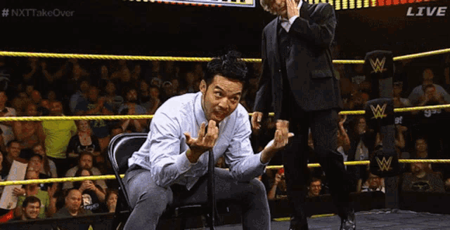 a man in a suit is sitting in a chair in a wrestling ring while another man stands behind him