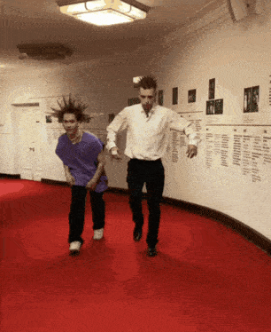 a man with a mohawk stands next to another man on a red carpeted hallway