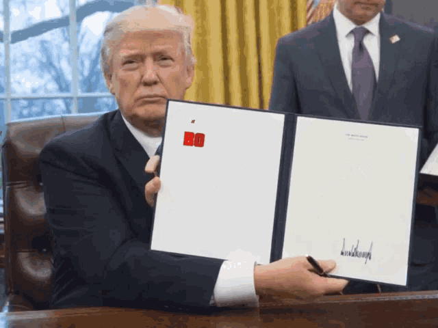 donald trump is sitting at a desk holding a document that says bo on it