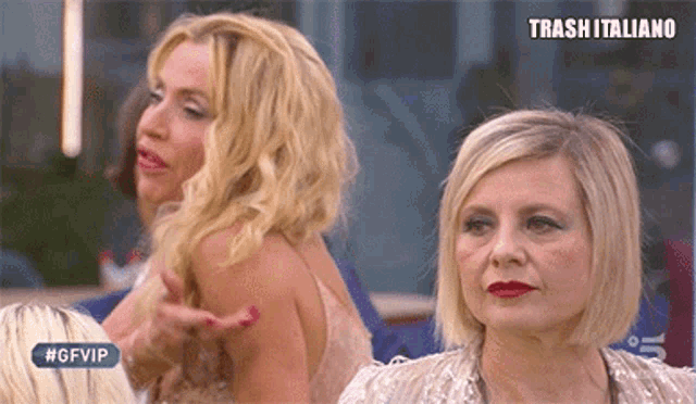 two blonde women are sitting next to each other in front of a sign that reads trash italiano