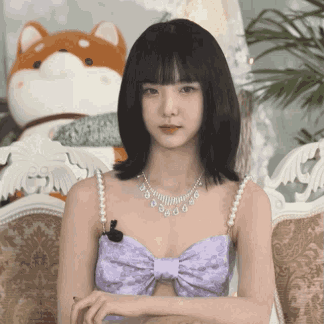a woman wearing a purple top with a bow and pearls is sitting on a couch
