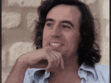 a man with long hair and braces on his teeth smiles with his hand on his chin