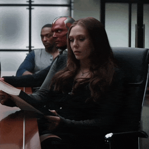 a woman sits at a table reading a book while a man in a red mask sits behind her