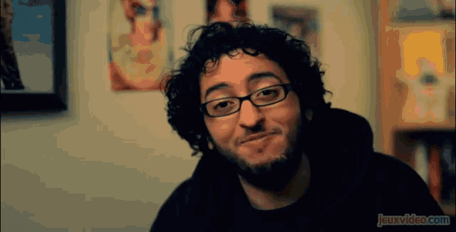 a man with glasses and a beard is smiling in front of a wall that says jeuxvideo.com on it