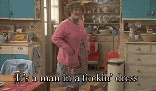 a woman in a pink sweater is standing in a kitchen with the words it 's a man in a fuckin ' dress