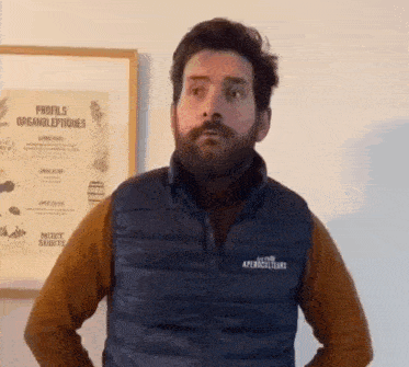 a man with a beard wearing a blue vest is standing in front of a wall with a poster on it .