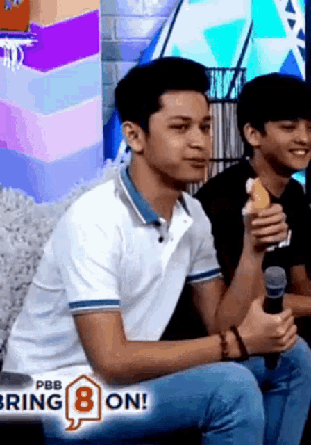 a man in a white shirt is holding a microphone and eating a banana while sitting next to another man .
