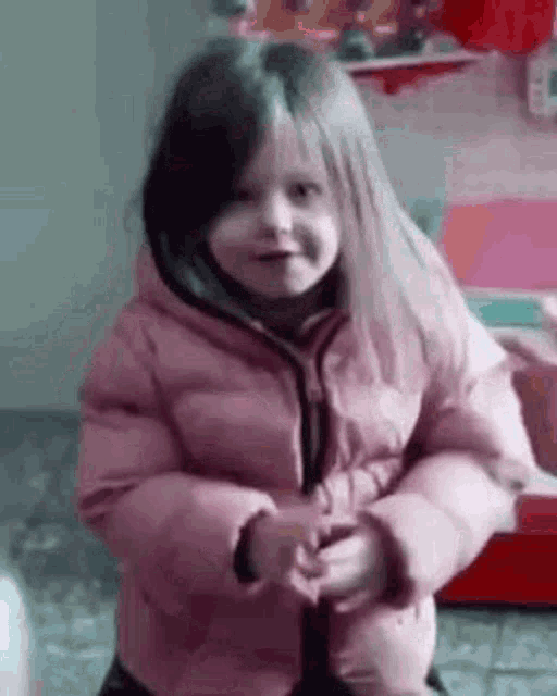 a little girl in a pink jacket is standing in a room and smiling .