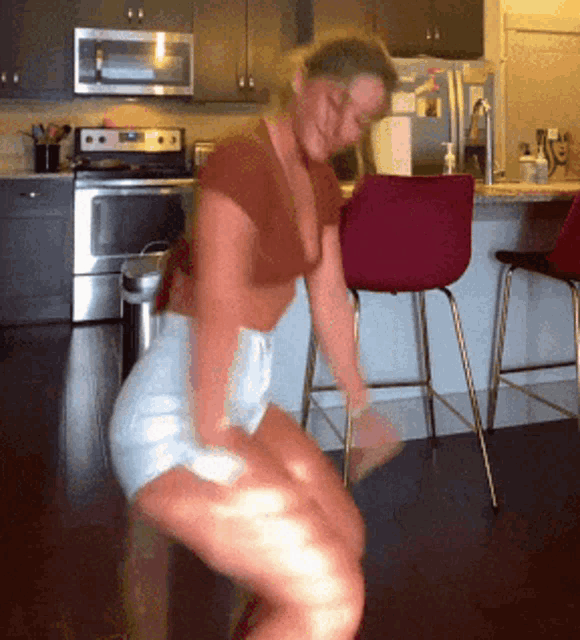 a woman in a crop top and shorts is squatting in a kitchen in front of a microwave