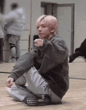 a young man with pink hair is sitting on the floor and pointing at the camera