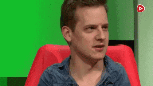 a man in a denim shirt is sitting in a red chair with a green screen behind him .