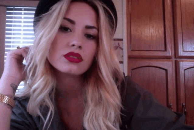 a blonde woman wearing red lipstick and a hat looks at the camera