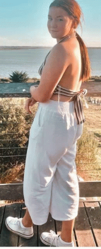 a woman in a bikini top and white pants is standing on a wooden deck overlooking the ocean .