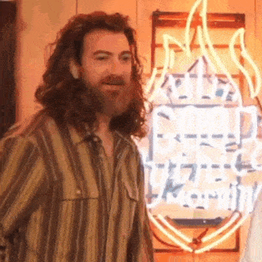 a man with long hair and a beard is smiling in front of a neon sign .