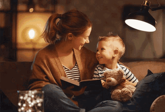 a woman sits on a couch reading a book to a young boy who is holding a teddy bear