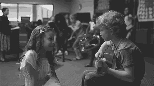a boy and a girl are sitting next to each other in a room . the boy is playing a guitar .