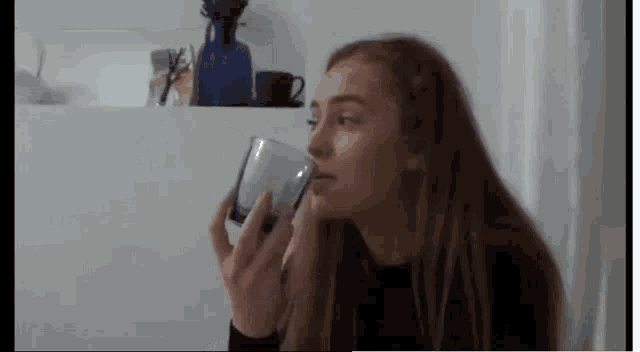 a woman is drinking from a glass in front of a shelf with vases on it .