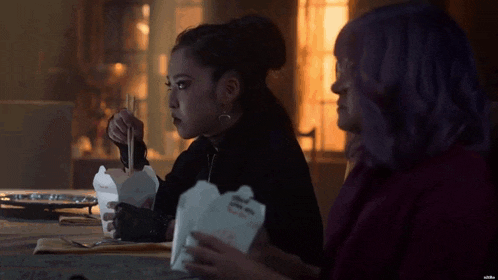 two women are sitting at a table eating noodles from a box with chopsticks