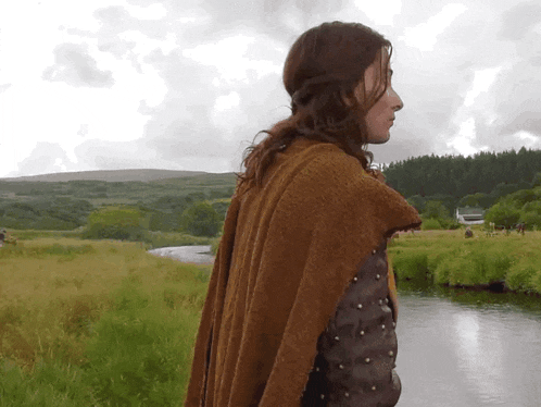 a woman in a brown cape stands in a grassy field near a river