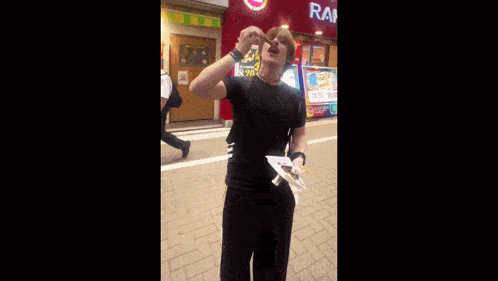 a man in a black shirt is eating a piece of food in front of a building that says ' rai ' on it