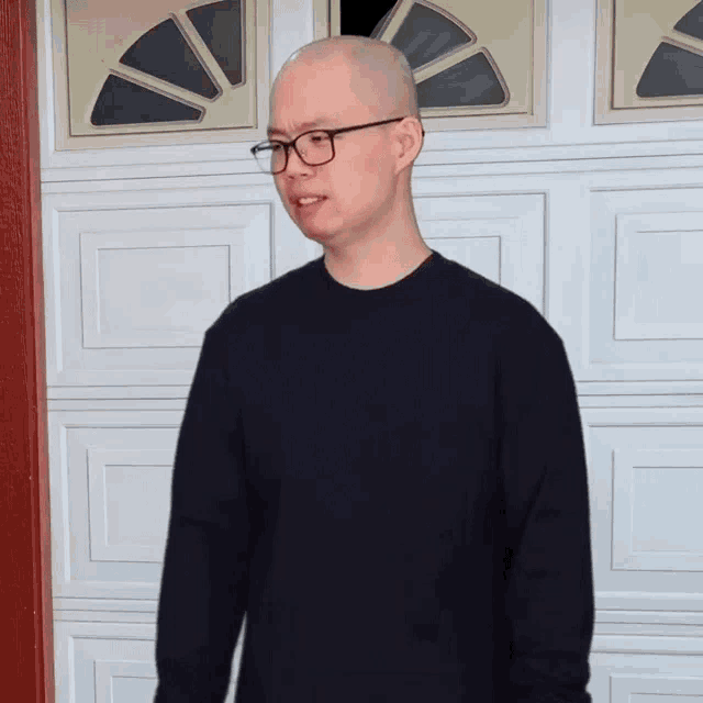 a bald man wearing glasses and a black sweater is standing in front of a garage door