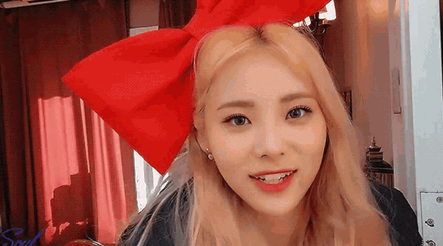 a blonde woman wearing a red bow on her head looks at the camera