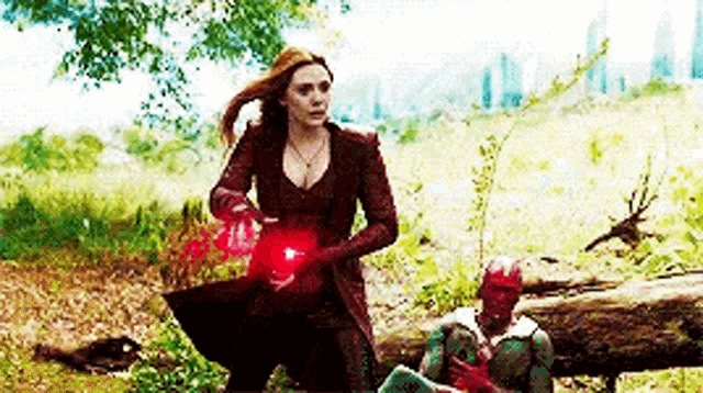 scarlet witch is holding a red sphere next to a man sitting on a log in a field .