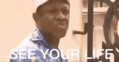 a man wearing a hat and a blue shirt is standing in front of a clock and says `` see your life '' .