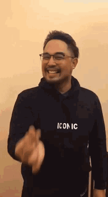 a man wearing a black hoodie with the word iconic on it is smiling and waving .
