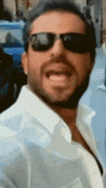 a man with a beard wearing sunglasses and a white shirt is smiling .