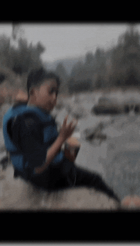 a boy wearing a life jacket sits on a rock by a river