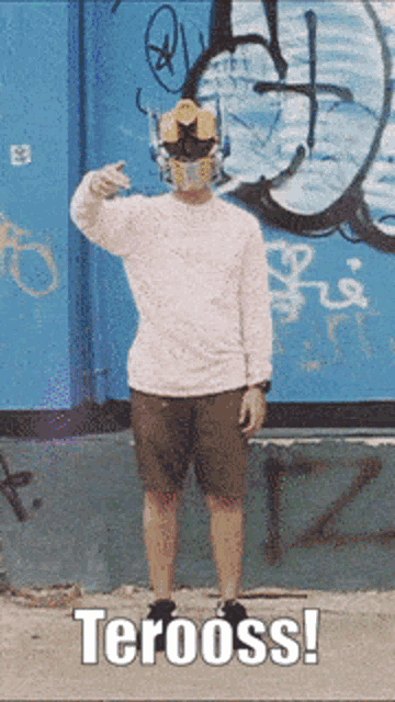 a man wearing a robot mask stands in front of a wall with graffiti and the words terooss