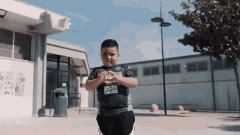 a young boy in a black shirt is standing in front of a building and making a heart shape with his hands .