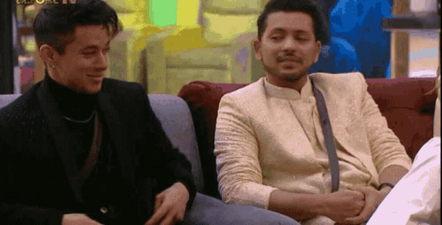two men are sitting on a couch and one of them is wearing a gold suit