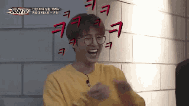 a young man wearing glasses and a yellow shirt is laughing while standing in front of a brick wall .