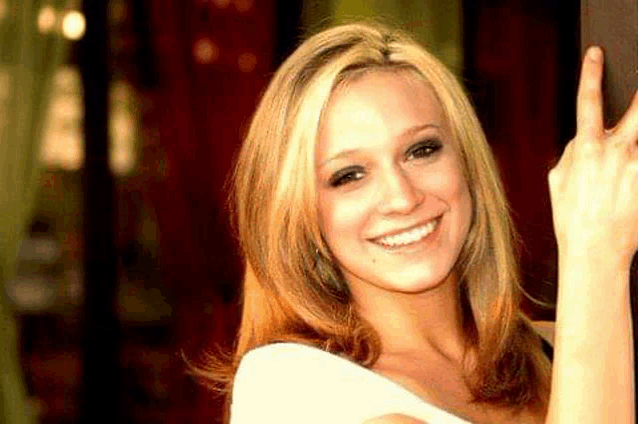 a woman is smiling and pointing at the camera while wearing a white shirt .